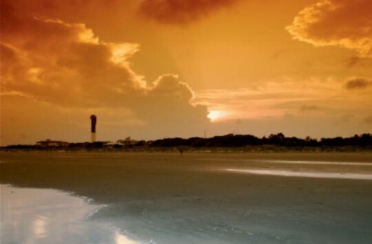 Sunset view of a beach on Sullivan’s Island, South Carolina, with a lighthouse in the distance and dramatic orange clouds reflecting on the wet sand.
